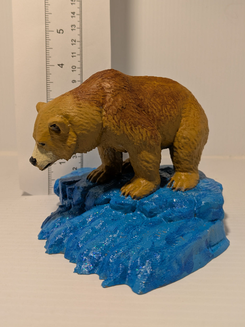 A small brown bear figurine standing on a blue icy base, photographed against a plain light background with a ruler standing behind it for scale.