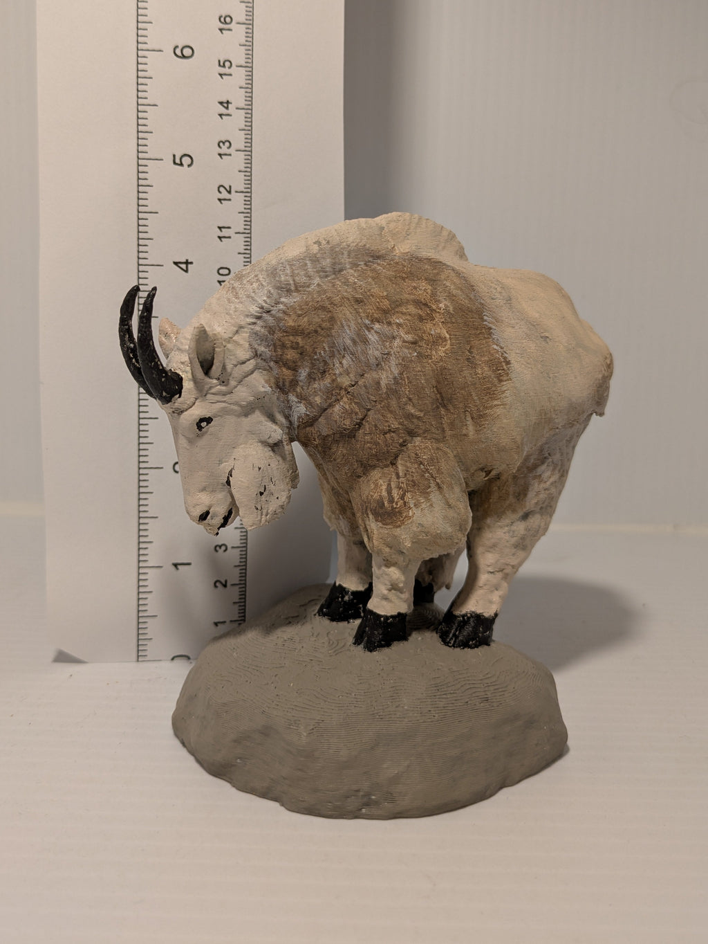 A small mountain goat figurine standing on a gray rock base, photographed indoors next to a white ruler for scale against a plain light background.
