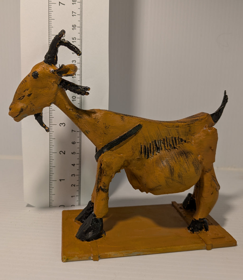 A hand-painted goat figurine in ochre yellow with black accents, standing on a matching rectangular base. A white ruler is positioned behind the figurine to show its height.