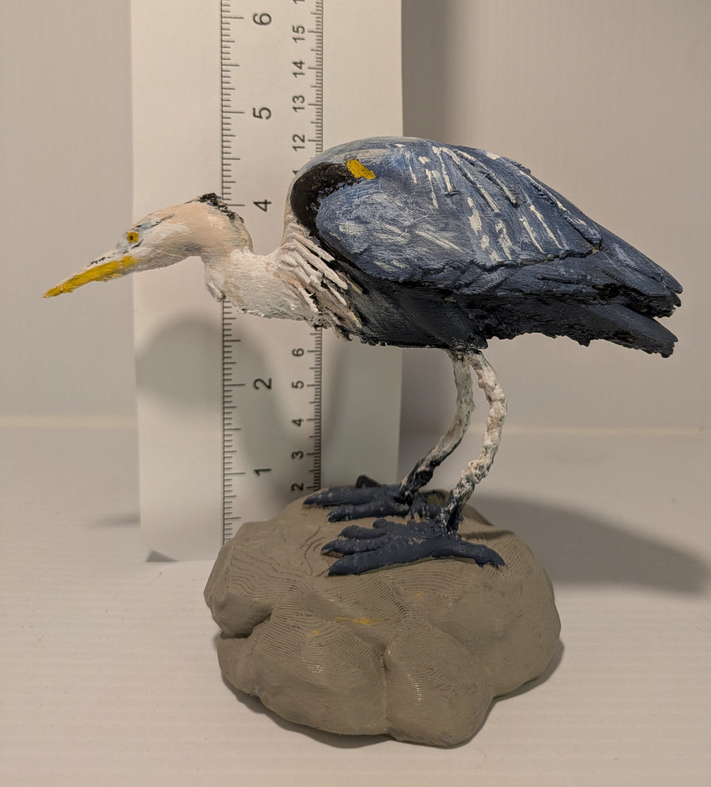 A handcrafted clay figurine of a heron standing on a rock base, shown next to a ruler for scale against a plain light background.